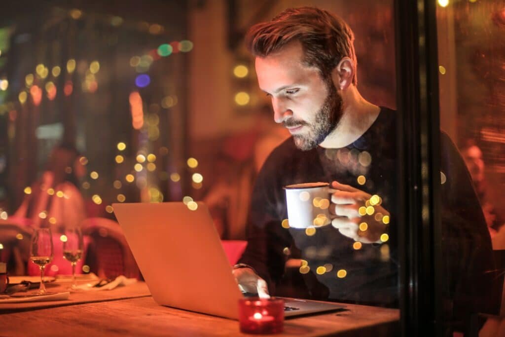 homem segura xícara de café, e está sob reflexo de luzes da janela, enquanto olha notebook